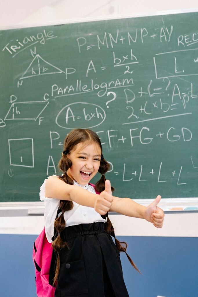 Cheerful girl in a classroom showing thumbs up with math formulas on the blackboard.